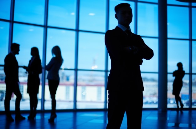 A silhouette of a confident businessman standing with arms crossed in the foreground of a modern office space with large windows. In the background, there are four other professionals, also in silhouette, engaged in conversation and collaboration.