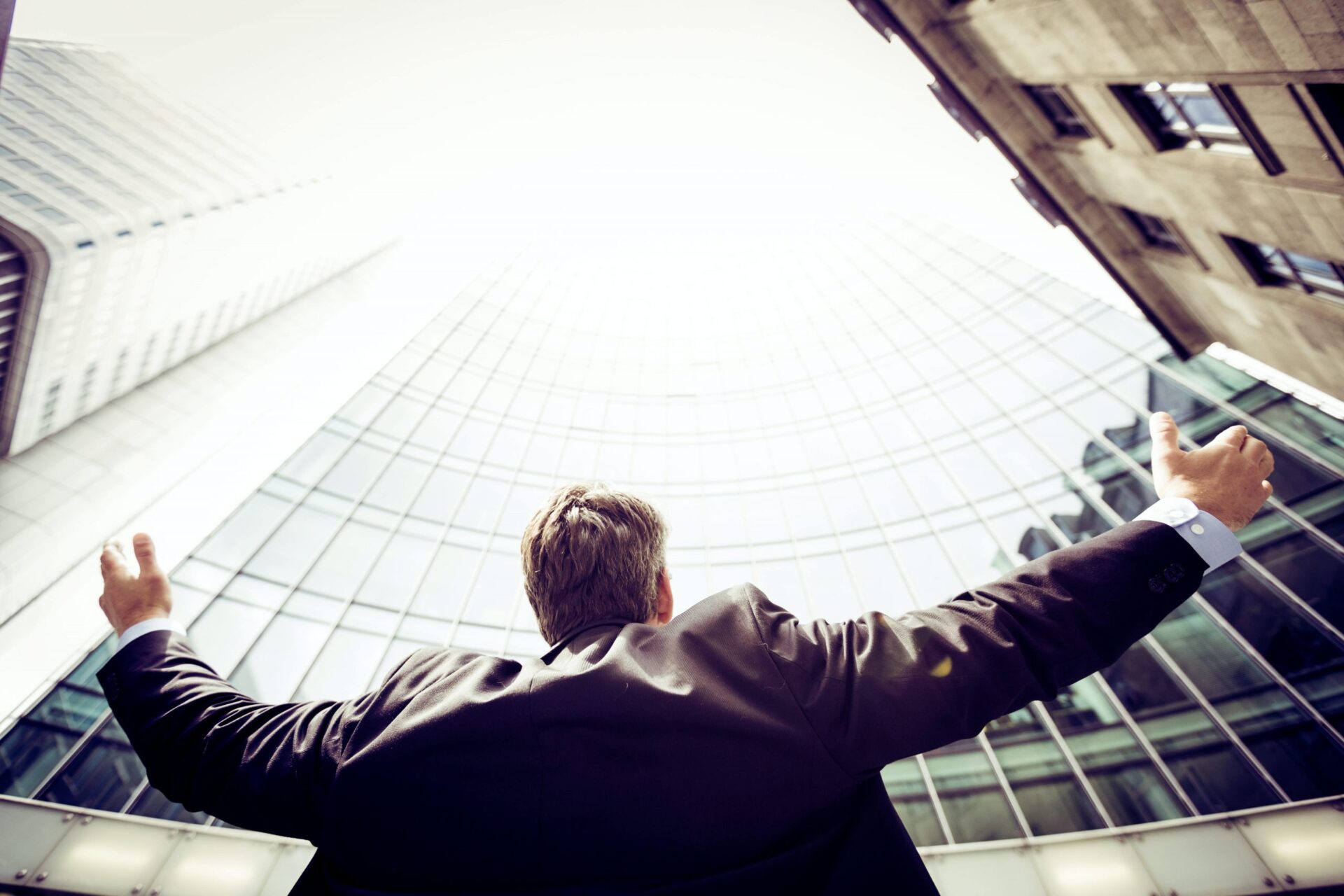 A low-angle photograph of a businessman standing with arms outstretched, looking up towards the sky in a modern cityscape. He is dressed in a suit and appears to be celebrating or feeling triumphant.