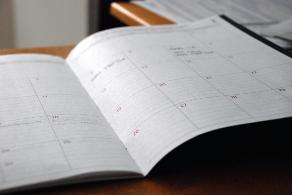 A close-up photograph of an open planner or calendar resting on a wooden desk. The pages show a monthly layout with dates and some handwritten notes and appointments. The focus is on the planner, with the background slightly blurred, hinting at other office items and papers nearby. The image conveys organization, planning, and the importance of keeping track of schedules and commitments.