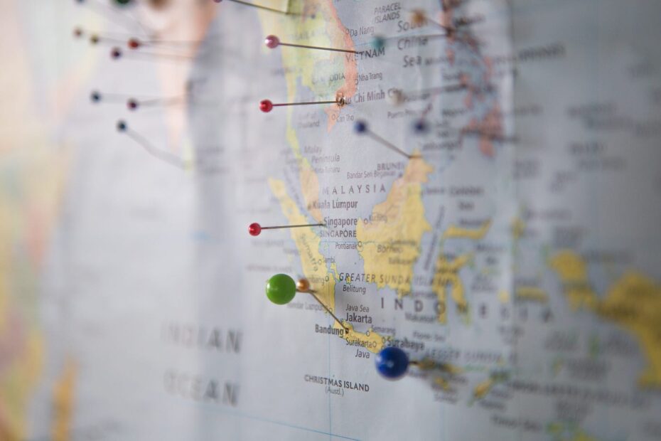 A close-up photograph of a world map with several colorful pins marking different locations. The pins, in various colors like red, green, and blue, are clustered around regions in Southeast Asia, including Malaysia and Indonesia. The focus is on the pins, with the map details slightly blurred, emphasizing the marked locations and suggesting themes of travel, planning, or tracking. The background is softly out of focus, keeping attention on the map and the pins.