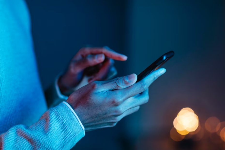 The image shows a close-up of a person using a smartphone, with both hands interacting with the device. The person is wearing a light-colored sweater, and the background is dimly lit with a soft, warm light in the distance, creating a cozy and intimate atmosphere. The focus is on the hands and the smartphone, emphasizing the act of texting, browsing, or engaging with digital content.