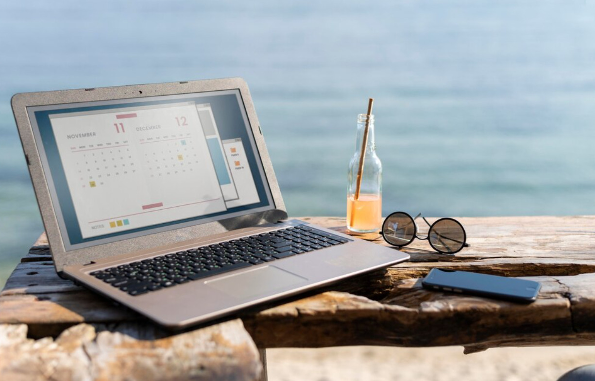 An image of a laptop and a sunglasses pair and bottle of orange juice at the beach.