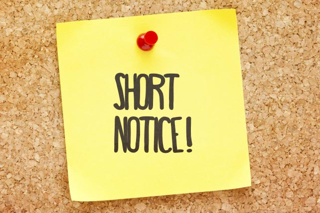 The image depicts a yellow sticky note pinned to a corkboard with a red pushpin. The note has the words "SHORT NOTICE!" written in bold, black letters, indicating urgency and a sudden announcement or change of plans. The corkboard background adds to the casual yet important nature of the message.