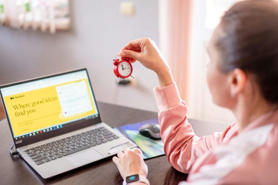 An image of a female employee sitting at her office desk, holding a tiny clock in her hand while working on her laptop, symbolizing time management and productivity.