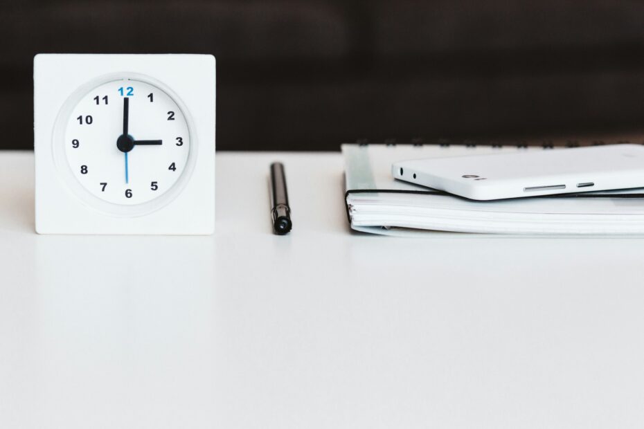 An image of a simple white disk featuring a small white clock, a notebook, and a pen arranged neatly on its surface, representing time management and organization tools.