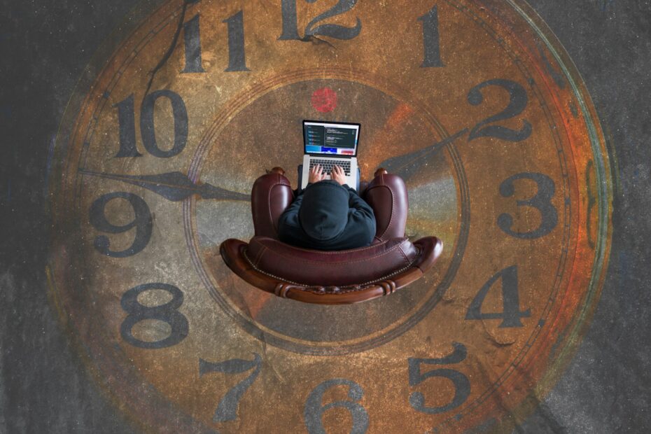 An image of an employee sitting at a desk, deeply focused on working on a laptop, with a large clock prominently displayed in the background, symbolizing time management