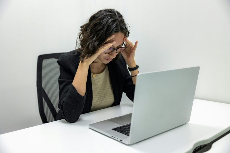 An image of a female employee sitting at her desk in front of a laptop, holding her head in her hands, appearing to have a headache or experiencing burnout. The setting suggests a stressful work environment.