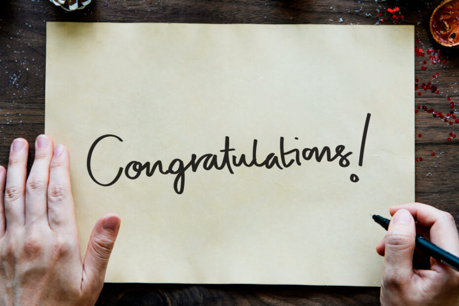 An image of a human hand holding a pen in front of a large piece of paper with the word "Congratulations!" written on it.