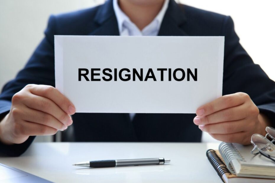 The image shows a person in a business suit holding a sign that reads "RESIGNATION." The individual is seated at a desk with a pen, notebook, and glasses, indicating a formal setting. The image clearly conveys the act of resigning from a job, emphasizing a significant and serious professional decision.