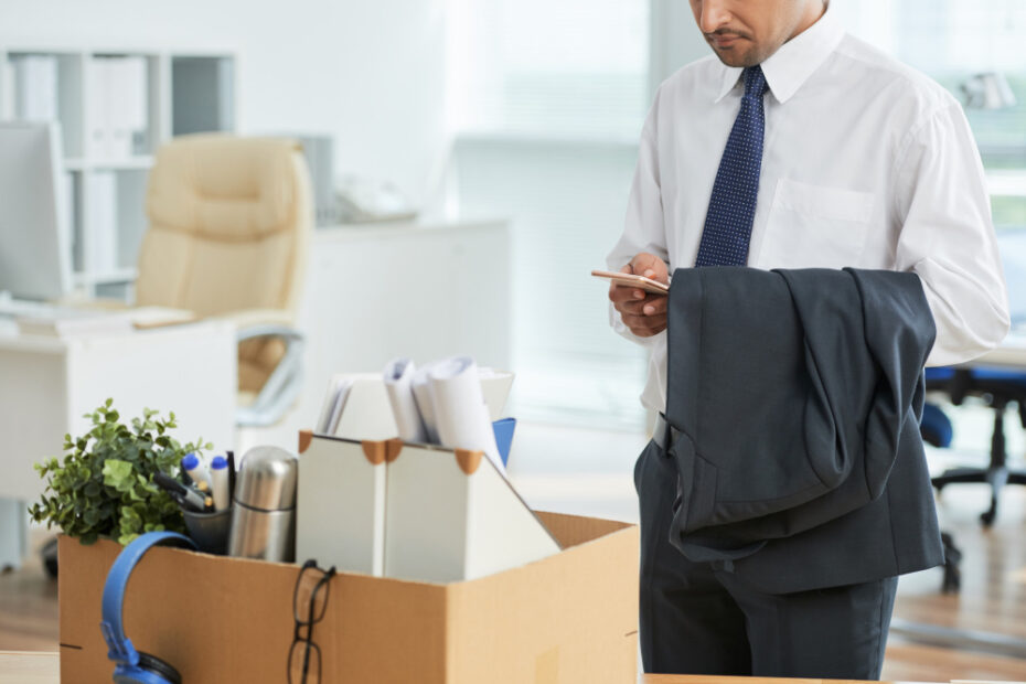 An image of an employee wearing a suit holding his phone, and standing in front of his disck with all his belongings in a cartoon box to leave his job after he quit