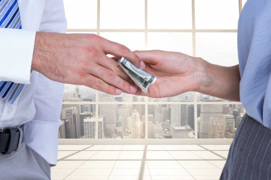 The image shows a close-up of two people exchanging money, with one person handing a rolled-up stack of cash to the other. Both individuals are dressed in business attire, indicating a professional setting. The background features large windows with a view of a city skyline, suggesting a high-rise office environment. The image conveys themes of financial transactions, business dealings, or potentially bribery.
