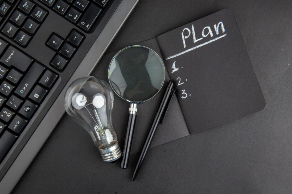 A black workspace features a black keyboard on the left and a black notepad labeled "Plan" with three numbered bullet points on the right. Surrounding the notepad are a magnifying glass, a lightbulb, and two pens, suggesting planning and brainstorming.