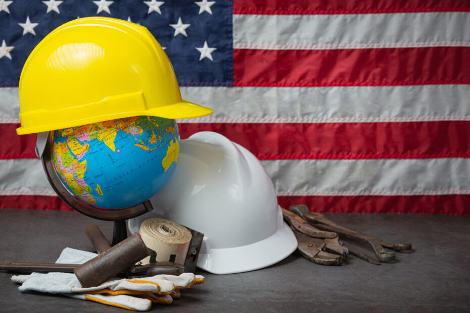 A globe wearing a yellow hard hat sits next to a white hard hat, various tools, and work gloves, all placed on a table. Behind them, the American flag is displayed, symbolizing labor and industry in the United States.