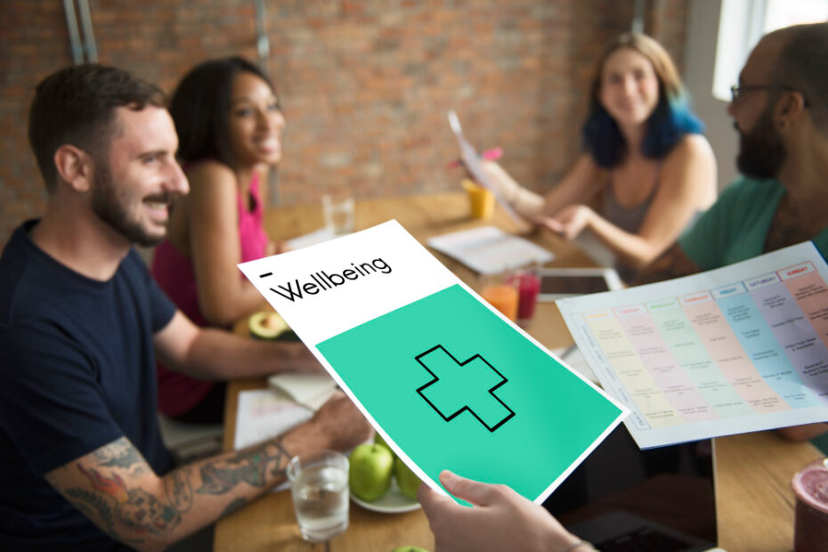 A group of colleagues sits around a table in a casual office setting, engaging in a discussion about well-being. One person holds a document with a green cross and the word "Wellbeing" prominently displayed. The team appears focused and engaged, with papers and healthy snacks like apples visible on the table.