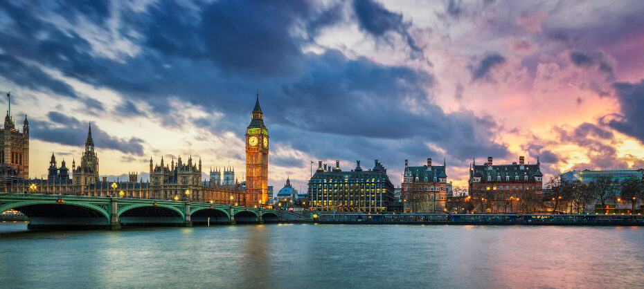 A panoramic view of London at dusk, showcasing the iconic Big Ben and the Houses of Parliament illuminated against a dramatic sky with a mix of dark clouds and vibrant hues of pink and purple. The River Thames reflects the lights from the buildings and the bridge, adding to the picturesque scene.