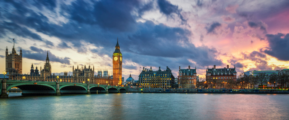 A panoramic view of London at dusk, showcasing the iconic Big Ben and the Houses of Parliament illuminated against a dramatic sky with a mix of dark clouds and vibrant hues of pink and purple. The River Thames reflects the lights from the buildings and the bridge, adding to the picturesque scene.