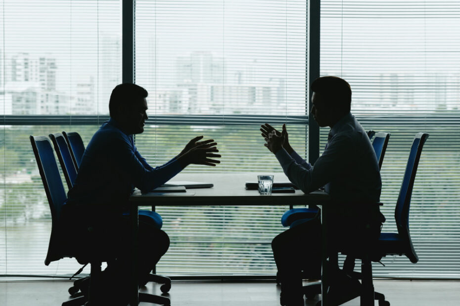 Two professionals are silhouetted against large windows during a discussion at a conference table in an office setting. The cityscape in the background is partially visible through the blinds. This image symbolizes a business negotiation or serious discussion.