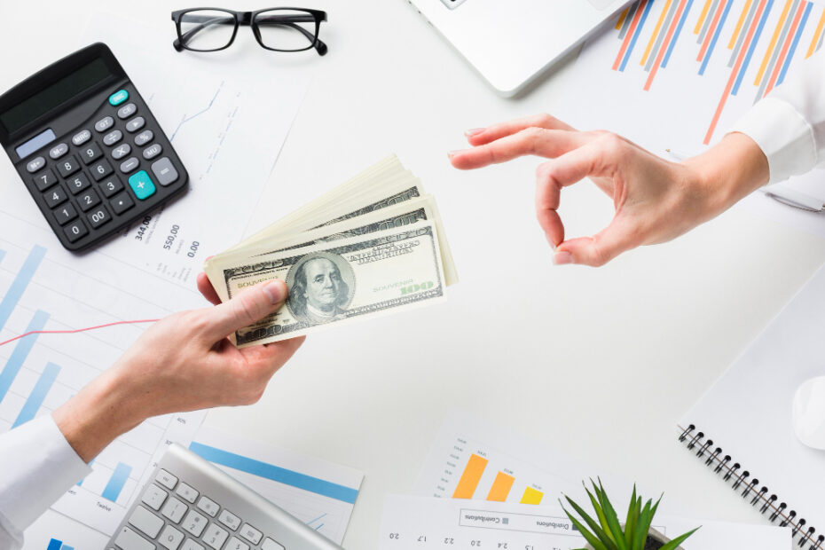 A person hands over a stack of hundred-dollar bills to another person making an "OK" gesture. The exchange takes place over a desk with financial charts, a calculator, glasses, a keyboard, and a small potted plant, symbolizing financial transactions or payroll.