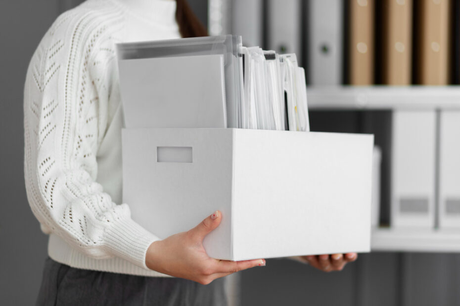A person in a white sweater holds a white box filled with neatly organized files, standing in front of shelves containing folders and binders.
