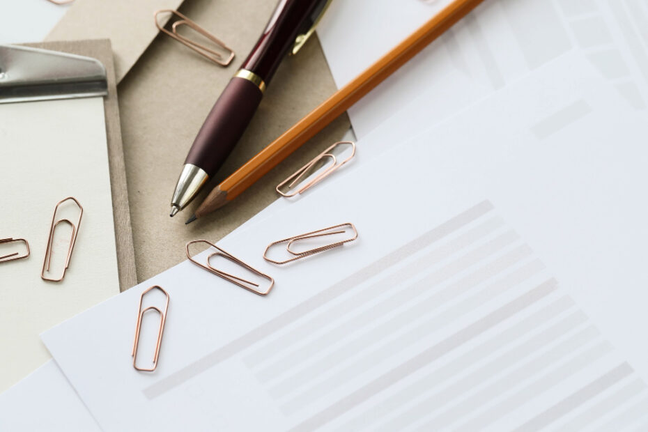 A close-up image of an office desk setup featuring a brown pen, a pencil, and several rose gold paper clips scattered on white sheets of paper. There is also a clipboard partially visible in the background, enhancing the professional and organized atmosphere.