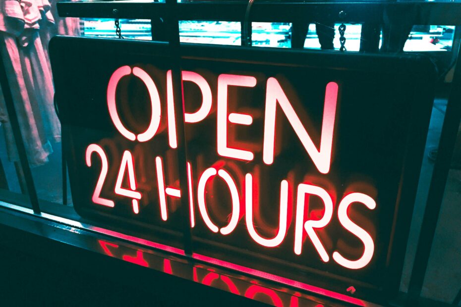 A brightly lit neon sign in a store window reads "OPEN 24 HOURS." The sign is glowing with red and white colors, indicating that the business operates around the clock. The background shows a dimly lit interior with faint outlines of people and store shelves.