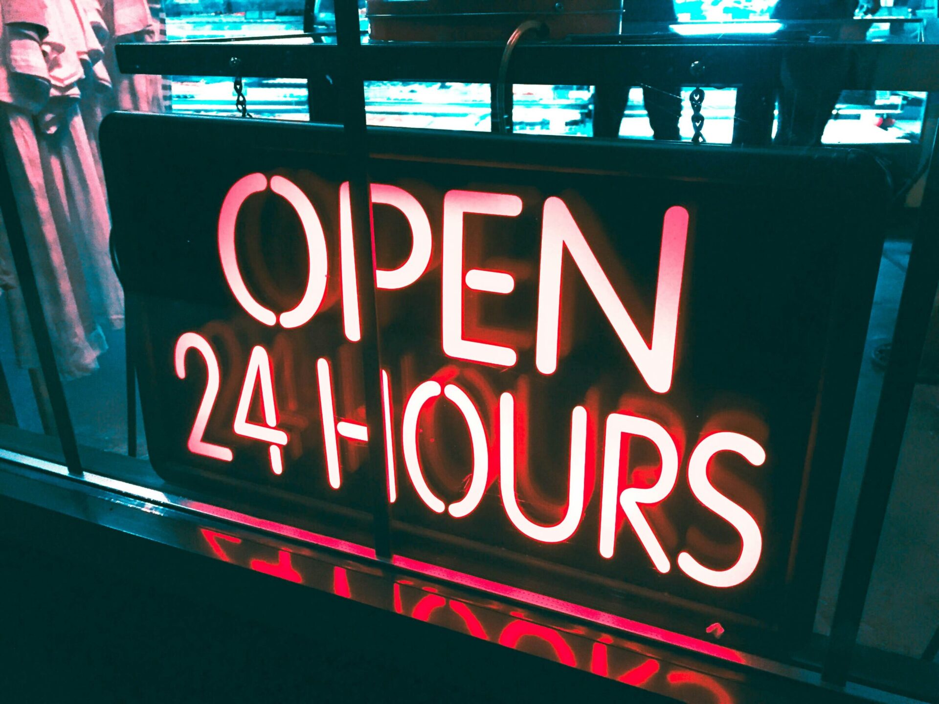 A brightly lit neon sign in a store window reads "OPEN 24 HOURS." The sign is glowing with red and white colors, indicating that the business operates around the clock. The background shows a dimly lit interior with faint outlines of people and store shelves.