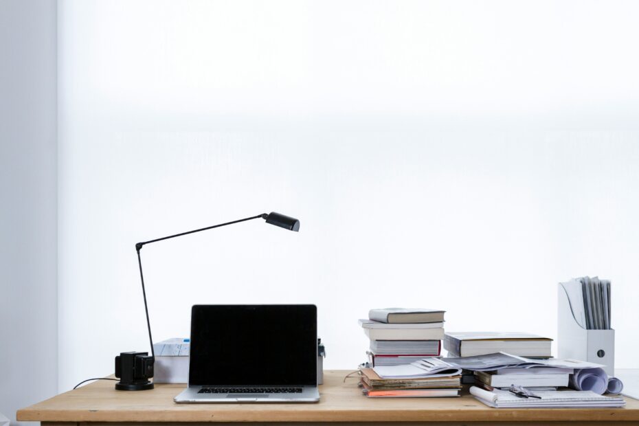 A minimalist office workspace with a wooden desk featuring an open laptop, a modern black desk lamp, and several stacks of books and documents on the right side. The background is a plain white wall, providing a clean and uncluttered environment.