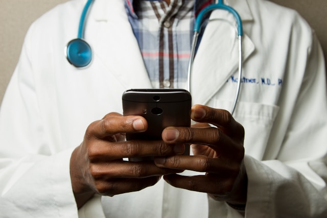 A doctor wearing a white lab coat and a stethoscope around his neck is holding a smartphone and typing a message. The image focuses on the doctor's hands and the phone, suggesting communication or checking medical information. The background is blurred, keeping the attention on the doctor's hands and the device.