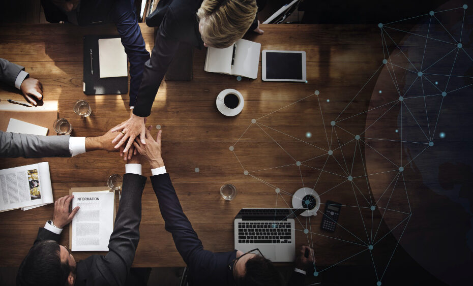 A group of business professionals is seated around a large wooden table, engaging in a collaborative meeting. The individuals are placing their hands together in a gesture of unity and teamwork, signifying collaboration and shared goals. The table is cluttered with open laptops, documents, notebooks, and cups of coffee, indicating a productive work session. In the background, a digital network overlay with connected dots and lines symbolizes global communication and interconnectedness in the business world.