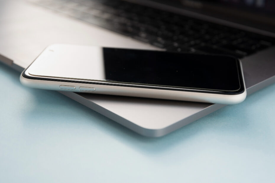 A close-up image of a smartphone resting on top of a closed laptop, both devices on a light blue surface. The smartphone screen is black, and the laptop's keyboard is partially visible in the background, creating a sleek, modern composition.