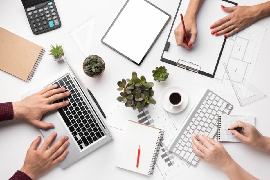 A top-down view of a collaborative workspace where multiple people are working together. The scene includes a laptop and a desktop keyboard being used, with one person typing on the laptop and another writing on a clipboard. There are notebooks, a calculator, a cup of coffee, plants, and various office supplies like rulers and pencils scattered around the desk. The workspace has a clean, organized look, with the plants adding a touch of greenery. The hands of the individuals suggest a teamwork environment, possibly in an office or a creative setting.