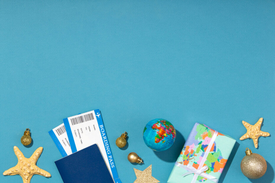 The image shows a travel-themed arrangement on a light blue background. It includes two boarding passes partially tucked into a blue passport, a small globe, a wrapped gift with a map pattern, and golden starfish ornaments alongside small gold Christmas baubles. The items suggest a holiday vacation, combining elements of travel with festive decorations, evoking the idea of celebrating the holidays while exploring new destinations.