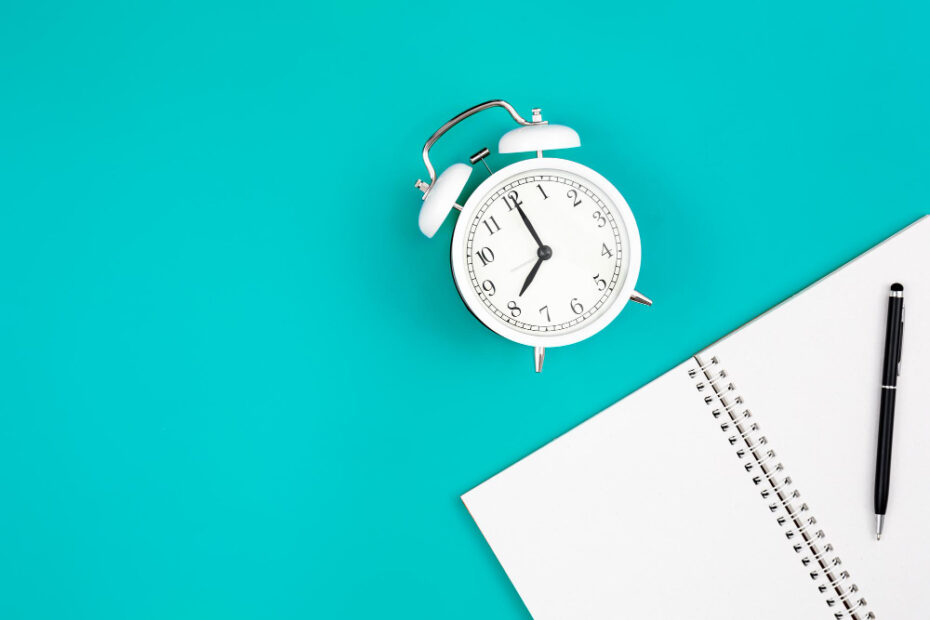 A minimalistic image featuring a white alarm clock on a vibrant turquoise background, symbolizing time management. To the right of the clock is an open spiral notebook with a black pen placed on top, suggesting planning or note-taking. The image conveys themes of organization, productivity, and efficient time usage.