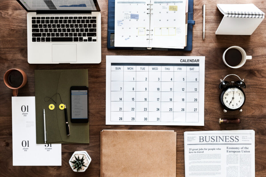 A well-organized workspace featuring a laptop, calendar, and various office supplies is shown from a top-down view. The desk includes a large printed calendar, a notebook, a smartphone, a cup of coffee, a clock, and a business newspaper. This setting reflects a professional environment focused on planning and managing tasks, including time off and scheduling, which aligns with the themes of transparent leave management and organized work culture