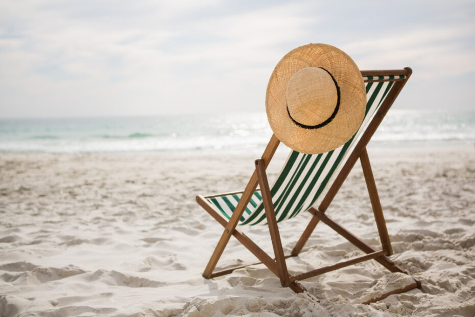 A beach scene featuring a striped deck chair with a straw hat draped over the backrest, facing the calm ocean. The sandy shore stretches out under a bright, partly cloudy sky, creating a peaceful and relaxing atmosphere perfect for a day of leisure by the sea