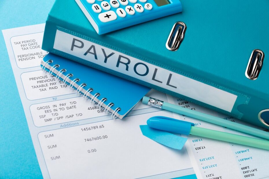 A blue binder labeled "PAYROLL" is placed on top of payroll documents, alongside a blue calculator, notebook, pen, and highlighter. The setup represents the organization and management of payroll processes in a business environment. The color-coordinated office supplies create a cohesive and professional workspace.