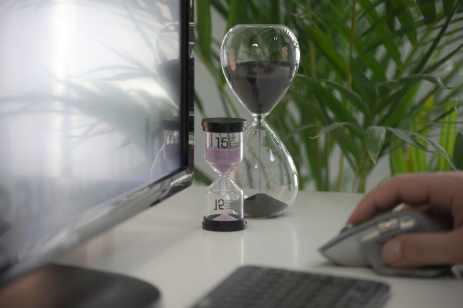 A close-up image of a work desk featuring two hourglasses next to a computer monitor. The larger hourglass contains black sand, while the smaller one, marked with the number "15," contains purple sand. A person's hand is visible on the right, using a computer mouse. The setup, complemented by green plants in the background, suggests a focus on time management or productivity in a work environment. The reflection of the hourglasses is visible on the screen, adding depth to the composition.