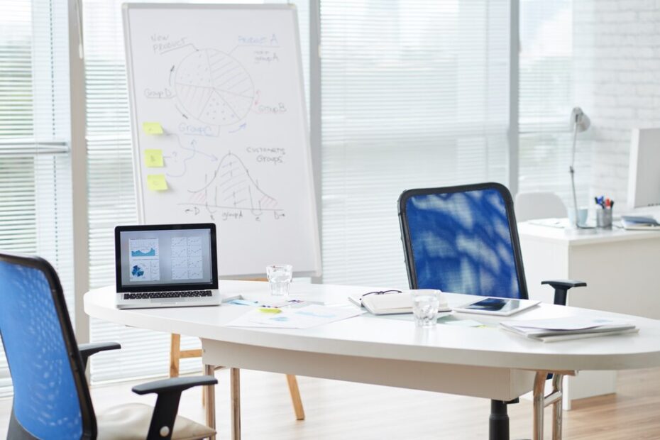 An organized office setting with a round desk featuring a laptop displaying charts, a notebook, and some documents. Two chairs face each other, indicating a meeting space. In the background, a whiteboard with graphs and sticky notes is visible, suggesting a discussion on business planning or data analysis. The atmosphere is professional and focused, reflecting a supportive work environment.