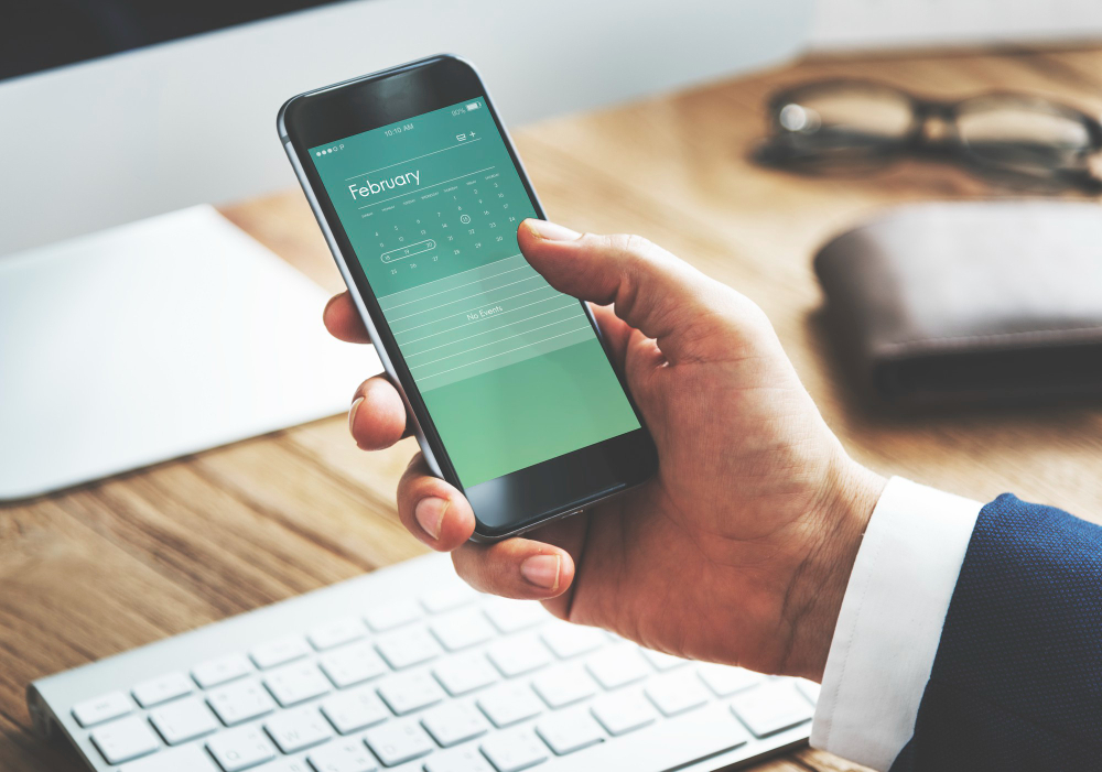 A close-up image of a person holding a smartphone in their hand, displaying a digital calendar for February. The calendar is likely part of an employee time off tracker app, showing the ease of managing leave requests on the go. The person is seated at a desk with a keyboard in the background, along with a wallet and glasses, suggesting a work environment. The image conveys the convenience and accessibility of using mobile technology for managing employee time off.