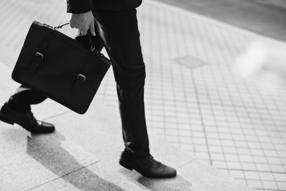 A black and white image of a person in business attire walking on a tiled sidewalk, carrying a leather briefcase in their left hand. Only the lower half of the person's body is visible, showing dress shoes and tailored trousers.