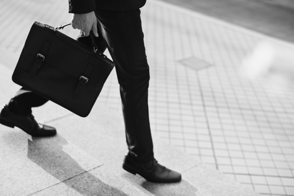 A black and white image of a person in business attire walking on a tiled sidewalk, carrying a leather briefcase in their left hand. Only the lower half of the person's body is visible, showing dress shoes and tailored trousers.