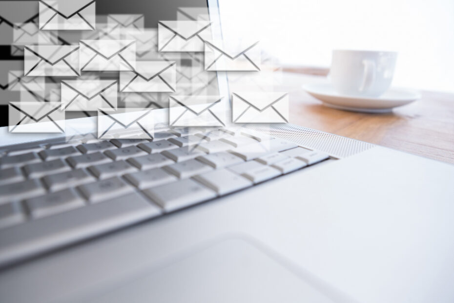 A laptop on a wooden desk, with numerous digital email icons floating above the keyboard, representing a flurry of email communication. A white coffee cup sits to the side, suggesting a work environment.