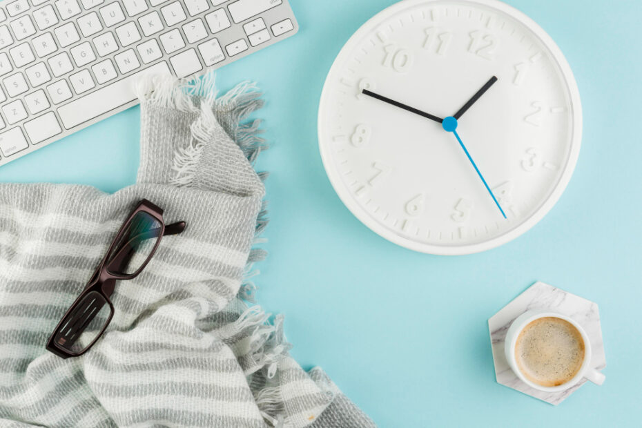 A minimalist workspace setup featuring a white keyboard, a large white wall clock showing the time, a cozy gray striped scarf, black eyeglasses, and a cup of coffee on a marble coaster, all arranged on a light blue background. This image conveys a relaxed yet productive atmosphere, ideal for managing time and tasks efficiently.