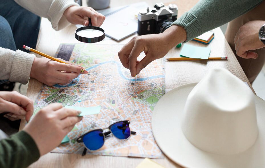 This image shows a group of people gathered around a table, planning a journey using a map. They are using pencils and a magnifying glass to examine the map details, indicating a careful and thorough planning process. A camera, sunglasses, and a stylish hat also appear, suggesting that they are preparing for a travel adventure.