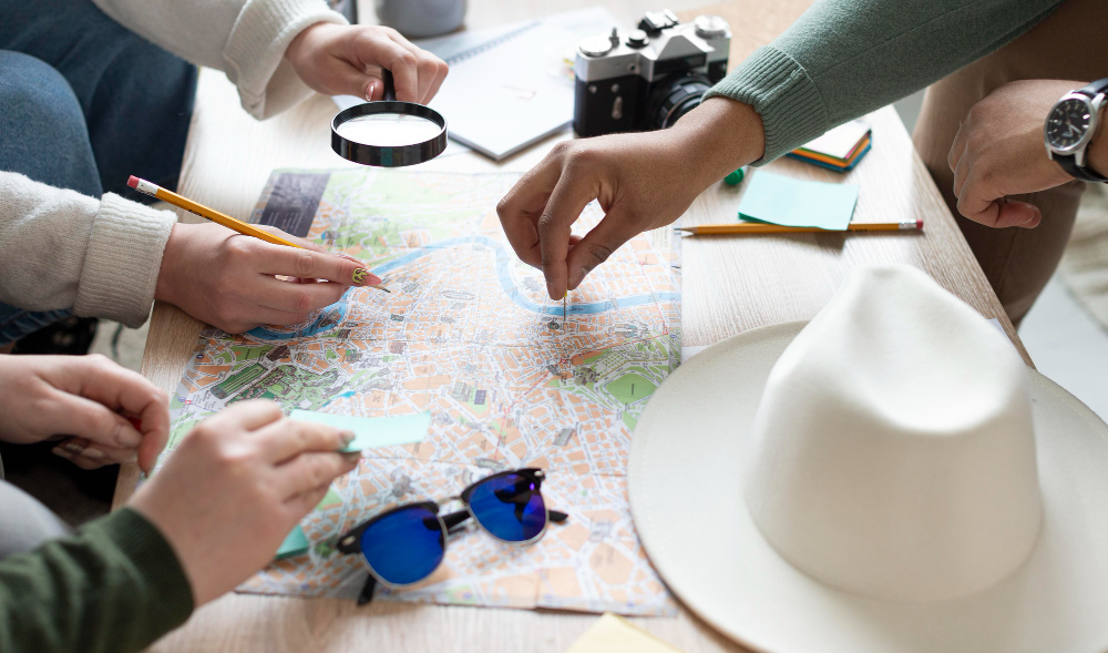 This image shows a group of people gathered around a table, planning a journey using a map. They are using pencils and a magnifying glass to examine the map details, indicating a careful and thorough planning process. A camera, sunglasses, and a stylish hat also appear, suggesting that they are preparing for a travel adventure.