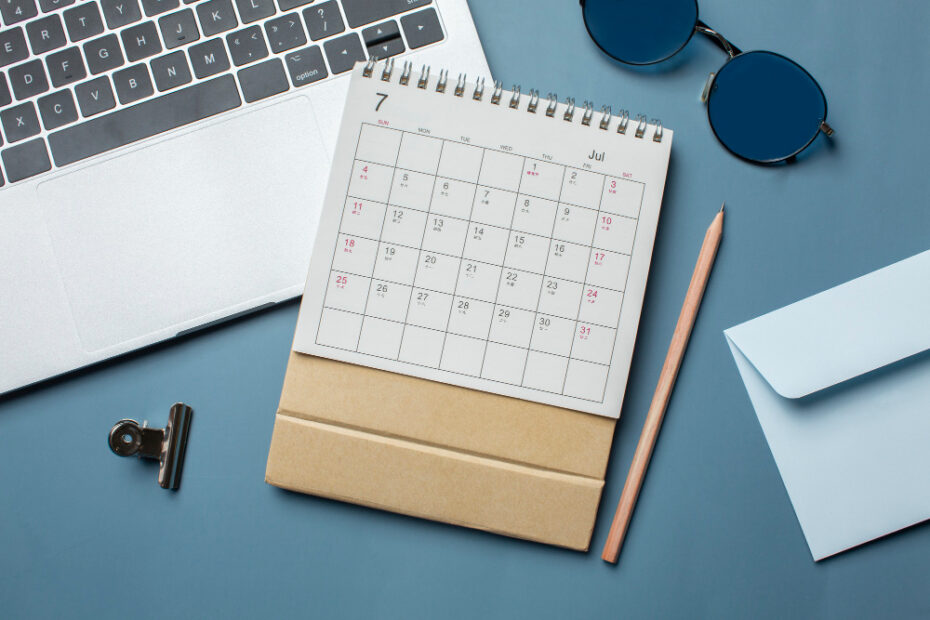 A close-up image of a desk setup featuring a calendar showing the month of July, a pencil, sunglasses, a laptop keyboard, a paperclip, and an envelope. The scene is organized on a blue surface, evoking a sense of planning, organization, and preparation for upcoming events or vacations.