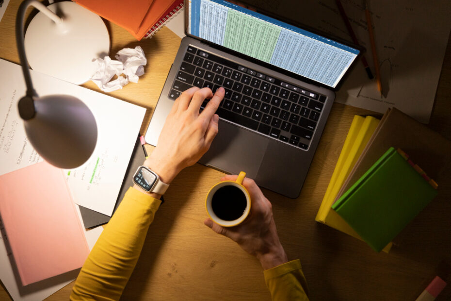 An overhead view of a person working at a desk with a laptop, holding a cup of coffee. The laptop screen displays a spreadsheet, suggesting the person is working on data analysis or financial tasks. Surrounding the laptop are various office supplies, including notebooks, papers, and a lamp, indicating a busy workspace. The warm lighting and casual setup create a focused, productive atmosphere.