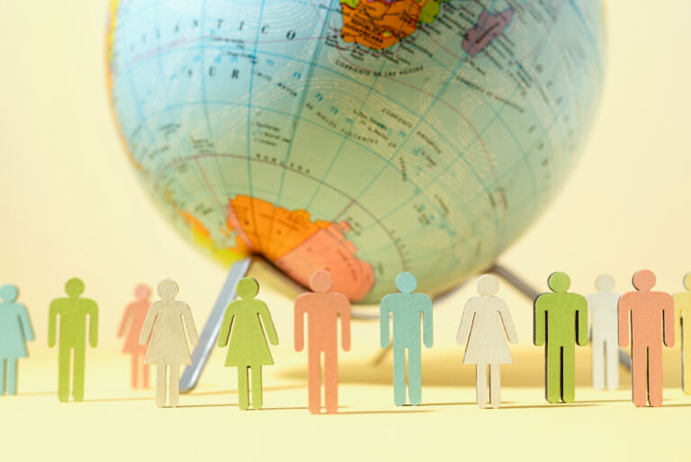 Paper cutout figures representing diverse people in various colors stand in a line in front of a globe, symbolizing global diversity and interconnectedness.