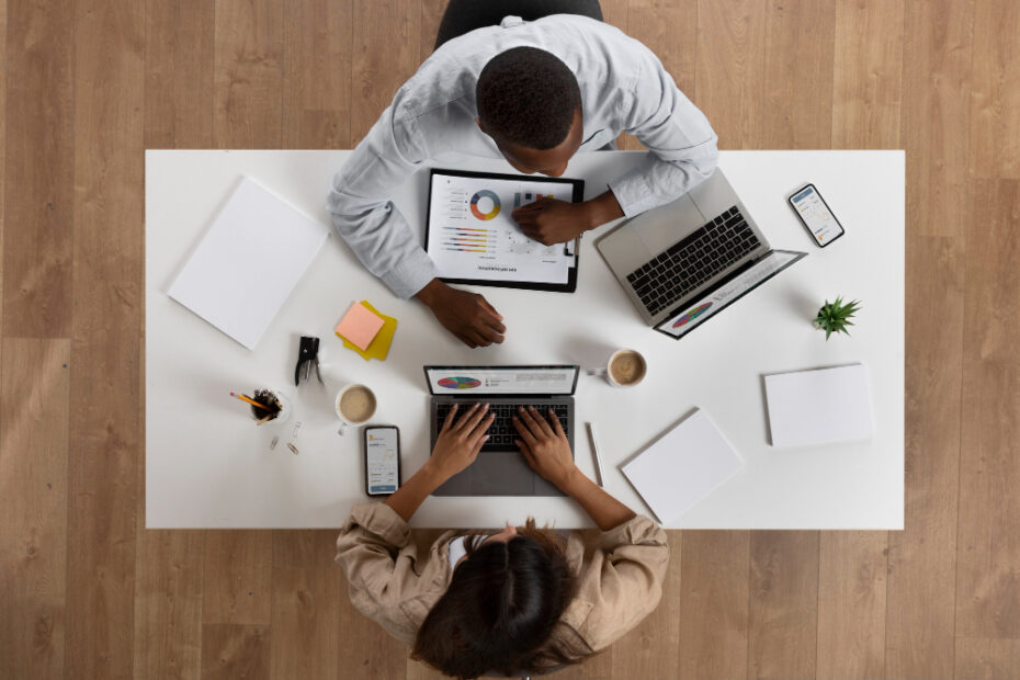 Two professionals are engaged in a collaborative meeting at a modern workspace. They are seated at a table equipped with various business tools: laptops, smartphones, printed documents with graphs, and notebooks. The setup indicates an organized and data-driven discussion, possibly involving planning, reviewing financial reports, or strategizing. Their focused interaction suggests they are analyzing information or making critical business decisions.