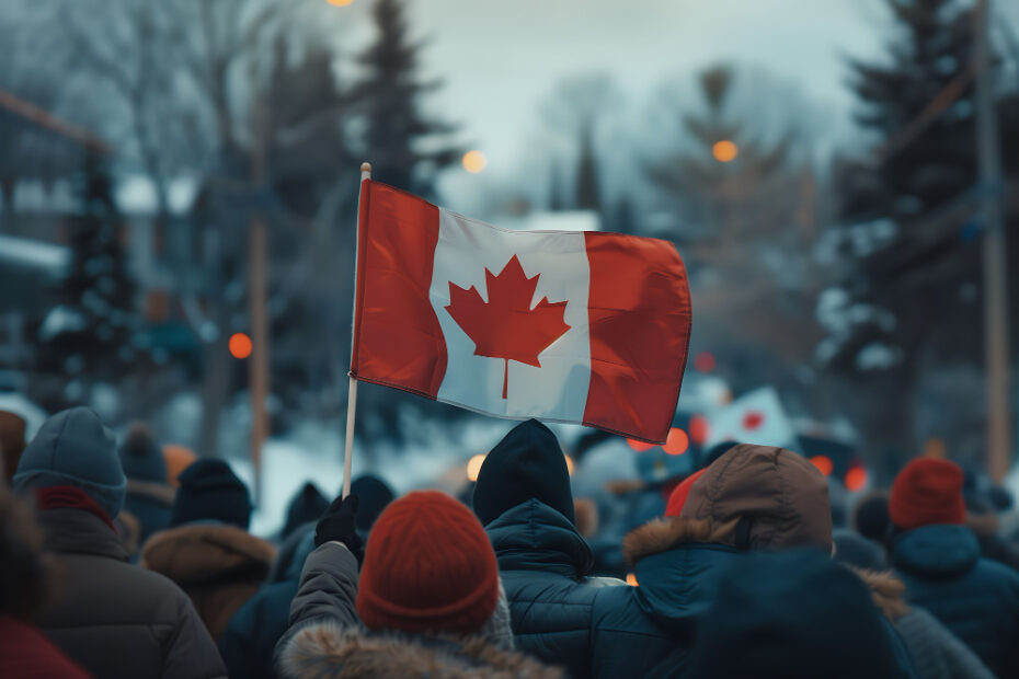 A group of people bundled in winter clothing gather outdoors, with one individual holding up a Canadian flag. The scene takes place in a cold, snowy setting with blurred trees and lights in the background, suggesting a national event or celebration during winter in Canada.